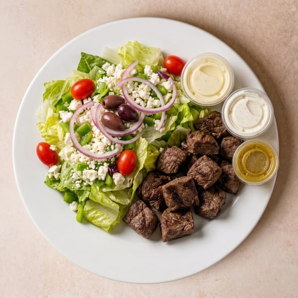 A white plate with a fresh salad, steak bites, and three small dressing cups.